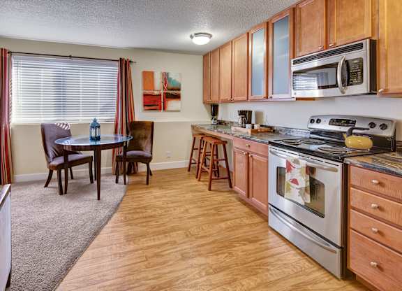 A bright open kitchen and dining space here at City View featuring warm wood cabinets, stainless steel appliances, a glass-top dining table with two chairs, striped curtains framing a large window, and wood-style flooring flowing into carpeted living space.