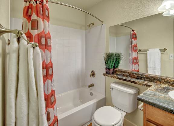 A bright bathroom here at City View featuring a white tub and shower with a curved rod and bold orange patterned curtain, a wide granite countertop with natural earth tones, and wood cabinetry below. A large mirror stretches across the wall, complemented by soft lighting, fluffy white towels, and a small faux plant for a touch of color.
