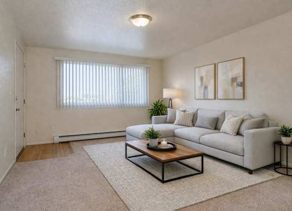 A bright living room here at Club at Eagle Pointe featuring a light gray sectional sofa with neutral pillows, a wooden coffee table on a soft area rug, and modern abstract wall art. A wide window with vertical blinds brings in natural light, creating an open, comfortable space ideal for everyday living.
