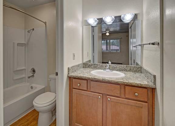 A clean and functional bathroom here at Club at Eagle Pointe featuring a granite-style vanity countertop with a single sink, warm wood cabinetry, and a large wall mirror with bright overhead lighting. The space includes a tub and shower combination with built-in shelving, durable flooring, and a layout designed for everyday comfort and convenience.