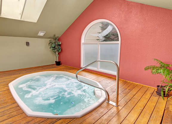 A relaxing indoor spa area here at Club at Eagle Pointe featuring a built-in hot tub with bubbling water, a wood-style deck surround, and a sturdy metal handrail for easy entry. The space is accented by warm-toned walls, potted greenery, and natural light from an arched window, creating a calm and inviting atmosphere.