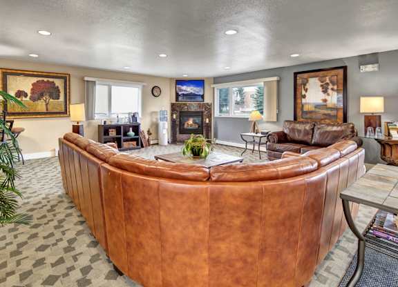 A spacious, inviting community lounge here at Club at Eagle Pointe featuring plush leather sectional seating, a cozy fireplace with a mounted TV, warm accent lighting, large windows for natural light, and tasteful artwork throughout, creating a comfortable gathering space for relaxing, socializing, or enjoying quiet time indoors.