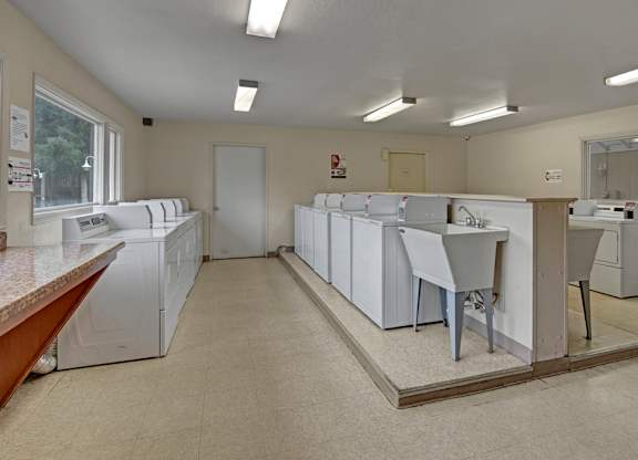 A clean, well-lit on-site laundry room here at Club at Eagle Pointe featuring multiple washers and dryers, a utility sink, wide walkways, bright overhead lighting, and durable flooring, providing residents with a convenient, functional space for everyday laundry needs.
