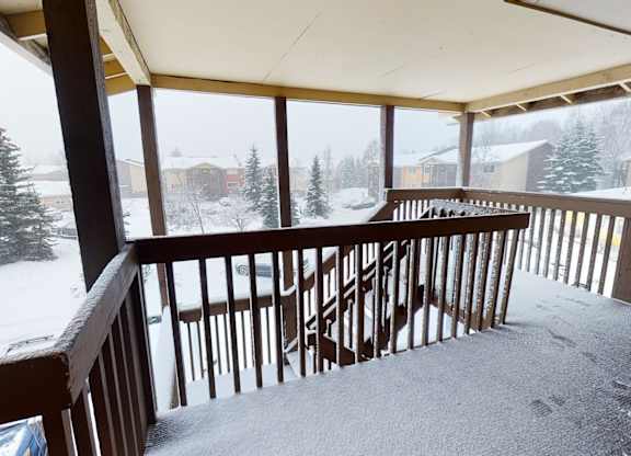 A covered outdoor walkway and stairwell here at Club at Eagle Pointe overlooking a snow-covered community courtyard, featuring wooden railings, upper-level access, and sheltered walkways that provide protected entry to apartment homes while showcasing a winter landscape with nearby buildings and evergreen trees.
