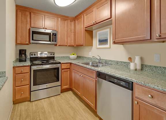 A warm, functional kitchen here at Club at Eagle Pointe featuring natural wood cabinetry, granite-style countertops, and stainless steel appliances including an oven, microwave, and dishwasher. Durable wood-style flooring and ample counter space create a practical layout that supports everyday cooking and comfortable meal preparation.