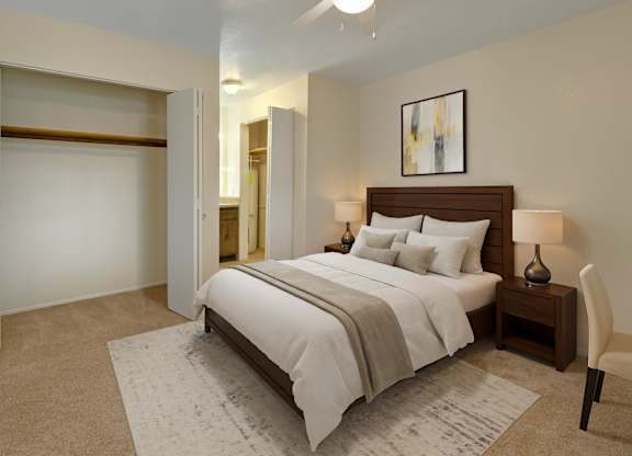 A spacious, inviting bedroom here at Club at Eagle Pointe featuring soft neutral walls, plush carpet flooring, and a comfortable bed with layered white bedding and accent pillows. An open closet with ample hanging space, bedside tables with lamps, and an adjacent bathroom create a functional, relaxing retreat designed for everyday comfort.