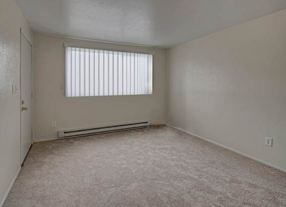 A bright, open bedroom here at Club at Eagle Pointe showcasing soft neutral walls, plush carpet flooring, and a wide window with vertical blinds that allow in natural light. A baseboard heater, simple layout, and generous floor space create a comfortable, versatile room ideal for relaxing, sleeping, or personalizing to suit your lifestyle.