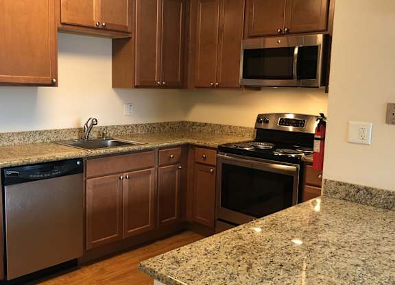 A spacious kitchen here at Club at Eagle Pointe featuring warm wood cabinetry, granite-style countertops, and stainless steel appliances including a refrigerator, range, microwave, and dishwasher. The functional L-shaped layout offers generous counter space, ample storage, and a breakfast bar ideal for meal prep and casual dining.