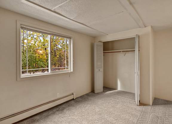 A comfortable bedroom here at College View featuring soft neutral walls, plush carpet flooring, and a large window that fills the space with natural light and views of mature trees outside. An open closet with shelving and hanging space adds practical storage, while the clean layout and baseboard heating create a cozy, functional retreat ideal for everyday living.