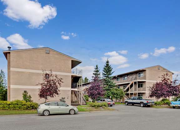 A welcoming exterior here at College View showcasing well-maintained apartment buildings with neutral siding, exterior staircases, and private balconies. Mature trees, landscaped greenery, and open parking areas create an inviting community setting, while clear walkways and open skies highlight the comfortable residential atmosphere.