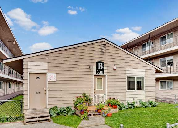 A charming exterior view here at College View featuring a well-kept community building with neutral siding, clearly marked entrances, and surrounding apartment structures with balconies. Lush green lawns, colorful potted plants, and tidy walkways create a welcoming residential atmosphere under bright blue skies.