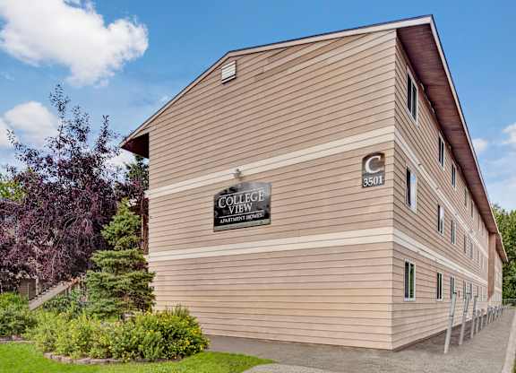 A welcoming exterior view here at College View highlighting a multi-story apartment building with neutral siding, large windows, and clearly marked building signage. Mature landscaping with trees and shrubs lines the walkway, creating a well-kept, residential setting under bright blue skies that reflects the community’s clean and approachable atmosphere.