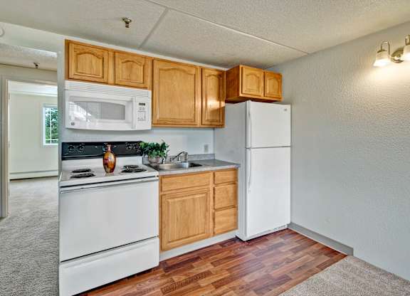 A cozy kitchen here at College View showcasing warm wood cabinetry, white appliances including a full-size refrigerator and range, and durable vinyl flooring that defines the cooking area. The functional layout offers ample cabinet storage, generous counter space, and a seamless connection to the adjacent living area, creating an efficient and comfortable everyday space.