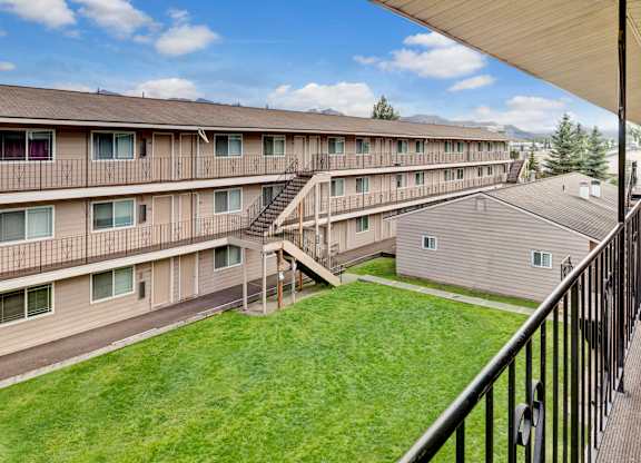A bright courtyard view here at College View showcasing multi-story apartment buildings with exterior walkways, railings, and staircases overlooking a well-maintained green lawn. Neutral siding, large windows, and open corridors create a clean, welcoming community layout with mountain views and blue skies visible in the background.
