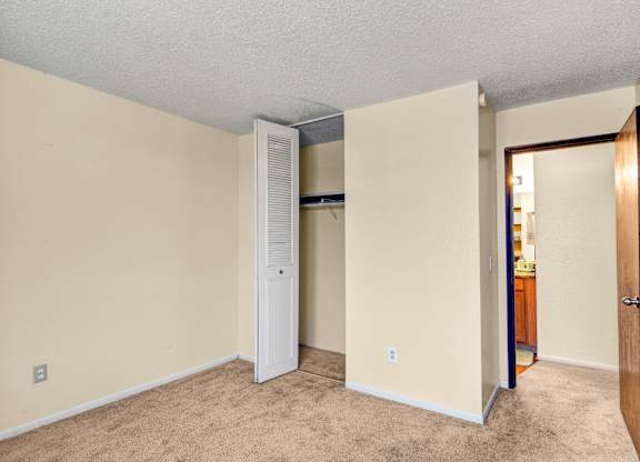 A bright bedroom here at Conifer Grove featuring soft carpet flooring, neutral walls, and a functional layout with an open closet and nearby bathroom access. The space offers a simple, flexible design with ample wall space for furniture, making it easy to create a comfortable and practical bedroom setup.