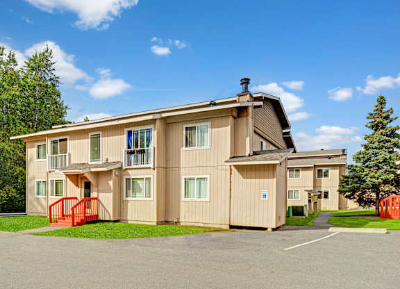 A bright exterior view here at Conifer Grove featuring a well-kept apartment building with neutral-toned siding, white-trimmed windows, and small private balconies, complemented by red entry railings, paved walkways, nearby parking, and surrounding greenery under a clear blue sky.