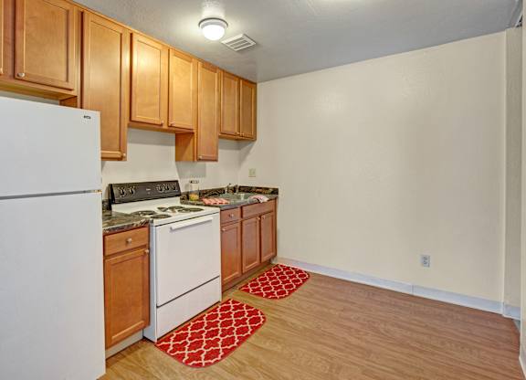 A functional kitchen here at Conifer Grove featuring warm wood cabinetry, laminate countertops, and a compact, efficient layout. White appliances include a full-size refrigerator, electric range, and sink, with wood-style flooring and overhead lighting providing a clean, practical space for everyday cooking.