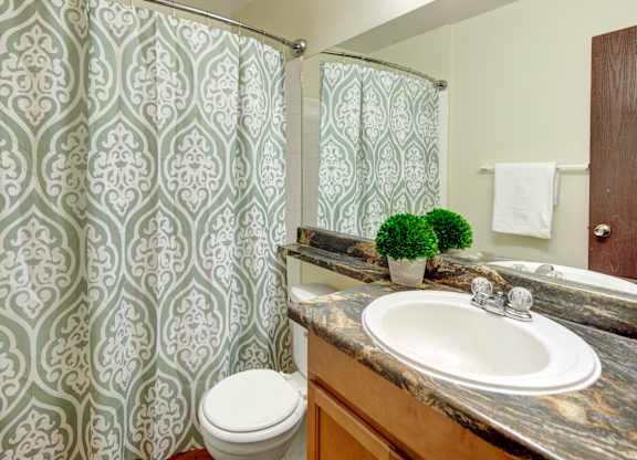 A fresh bathroom here at Conifer Grove featuring a clean tub and shower combination with a patterned shower curtain, a spacious vanity with wood cabinetry, and a durable countertop with an integrated sink. The space includes a large wall mirror, neutral wall tones, and simple fixtures that create a functional and comfortable daily routine.