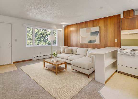 A bright living room here at Country Lane featuring a light neutral sectional sofa, wooden coffee table, and beige area rug, with a wood-paneled accent wall, large window providing natural light, and an open kitchen pass-through creating a warm, inviting apartment layout.