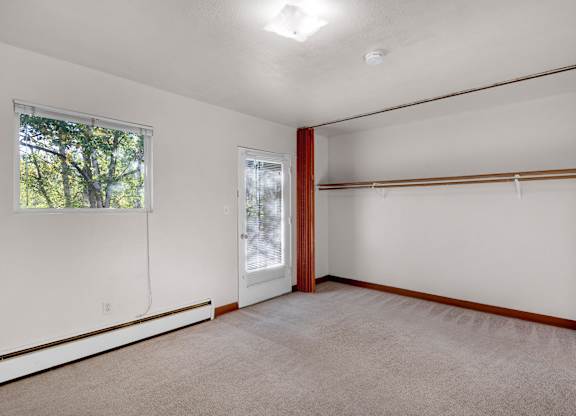A spacious bedroom here at Country Lane featuring neutral white walls, plush carpet flooring, warm wood trim, an open wall-length closet with shelving and hanging rods, and a glass door leading outside that brings in natural light and enhances everyday functionality.