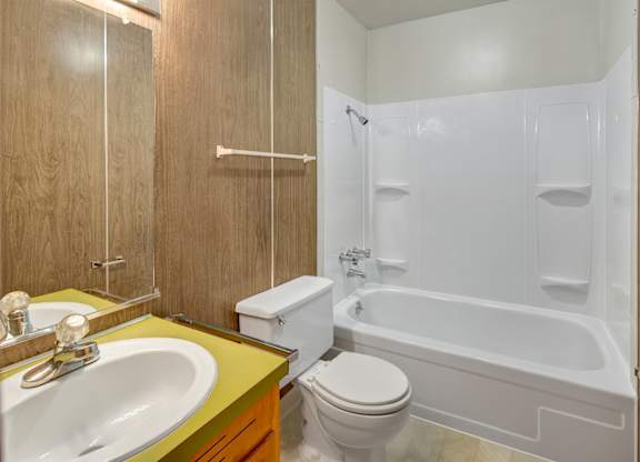 A clean bathroom here at Country Lane featuring a white tub and shower surround with built-in shelving, a single-sink vanity with a yellow countertop, a mirrored medicine cabinet, wood-paneled walls, and durable flooring designed for easy everyday use.