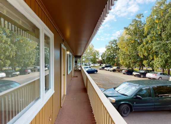 A covered exterior walkway here at Country Lane featuring tan siding, a private entry door, wide windows, and railing-lined views overlooking the parking area and surrounding mature trees for an open, airy feel.