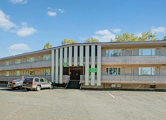 A welcoming exterior view here at Country Lane featuring a two-story apartment building with covered walkways, central stair access, private entry doors, large windows, and convenient on-site parking set among mature trees under a clear blue sky.
