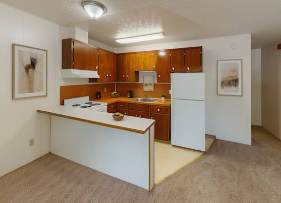 A functional kitchen here at Country Lane featuring warm wood cabinetry, white appliances, and an open breakfast bar overlooking the living area, with neutral flooring, simple wall art, and bright overhead lighting creating a clean and practical apartment layout.