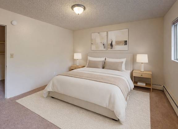 A bright bedroom here at Country Lane featuring a queen-sized bed with white bedding and neutral accent pillows, matching nightstands with lamps, soft carpeting, an area rug under the bed, abstract wall art, and an open closet providing simple storage and a calm, comfortable layout.