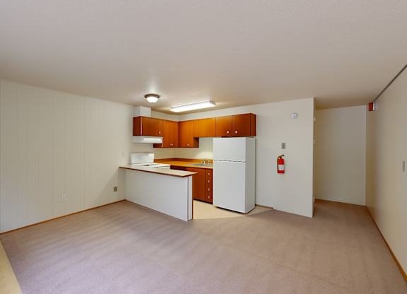A spacious living area here at Country Lane showcasing soft carpet flooring, neutral walls, and an open layout that flows into the kitchen, which features warm wood cabinetry, white appliances, ample counter space, and overhead lighting that creates a functional and inviting everyday living space.