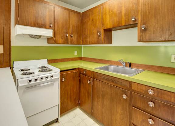 A functional kitchen here at Country Lane featuring warm wood cabinetry, bright green countertops, white appliances, a stainless steel sink, and ample cabinet storage, offering a practical layout with durable finishes and easy access to everyday cooking and meal preparation.