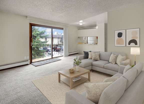 A bright living room here at Deer Park featuring a spacious open layout with a light gray sectional sofa, neutral throw pillows, a wooden coffee table, and soft area rug, complemented by modern wall art, a floor lamp, and sliding glass doors that open to a private balcony with outdoor views.