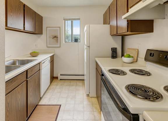 A bright kitchen here at Deer Park featuring warm wood cabinetry, white countertops, a double sink, dishwasher, electric range, and refrigerator, with a compact galley layout, vinyl flooring, neutral wall art, and a window that brings in natural light.