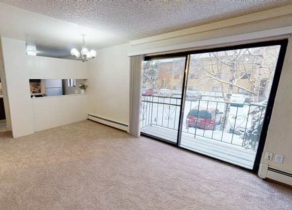Bright living room here at Deer Park featuring wall-to-wall carpet, neutral walls, and a spacious open layout. Sliding glass doors lead to a private balcony, allowing natural light and outdoor views, while a passthrough window connects the living area to the kitchen for added functionality.