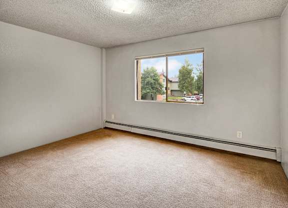 Airy bedroom here at Deer Park featuring wall-to-wall carpet, light neutral walls, and a large window that brings in natural light and outdoor views. A simple layout provides flexible space for bedroom furniture, while baseboard heating and an overhead light add everyday comfort and functionality.