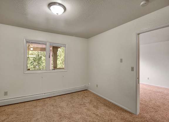 A bright bedroom here at Driftwood featuring plush neutral carpeting, light walls, and a large window that brings in natural light and leafy views, with a comfortable layout that connects easily to an adjacent room, creating a flexible space ideal for sleeping, relaxing, or setting up a home office.