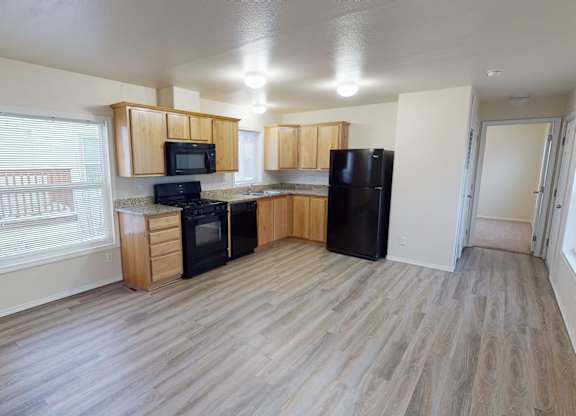 A bright open kitchen here at Four Seasons featuring light wood cabinetry, granite-style countertops, and black appliances that create a clean, modern look. Wide plank flooring flows throughout the space, while large windows and ceiling lights provide ample natural and overhead lighting, connecting seamlessly to the adjacent living area and hallway.