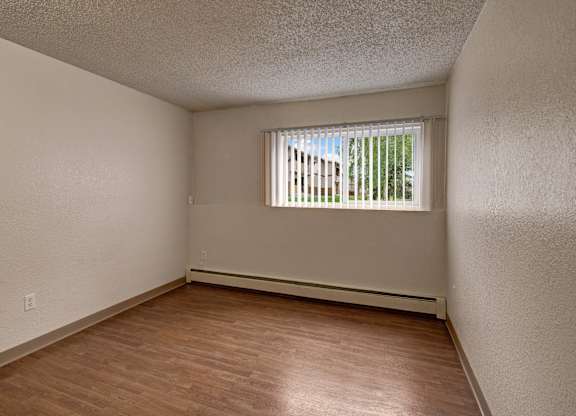 A bright unfurnished bedroom here at The Greenbriar featuring neutral painted walls, wood-style flooring, and a large window with vertical blinds that allows natural light and outdoor views. The room includes baseboard heating, clean lines, and an open layout that offers flexibility for bedroom furniture placement.