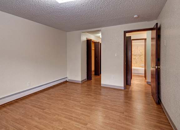 A spacious unfurnished bedroom here at The Greenbriar featuring neutral painted walls, wood-style flooring, and an open layout with clear access to a hallway, closet area, and adjacent bathroom. The room includes baseboard heating, a textured ceiling with overhead lighting, and ample wall space that allows flexible furniture placement and comfortable everyday living.