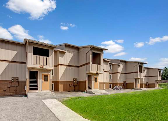 A bright exterior view here at The Greenbriar featuring a row of two-story apartment buildings with neutral tan and brown siding, private covered balconies, and clearly marked entrances. The image highlights well-kept green lawns, concrete walkways, and convenient access points, all set beneath a clear blue sky with soft white clouds.