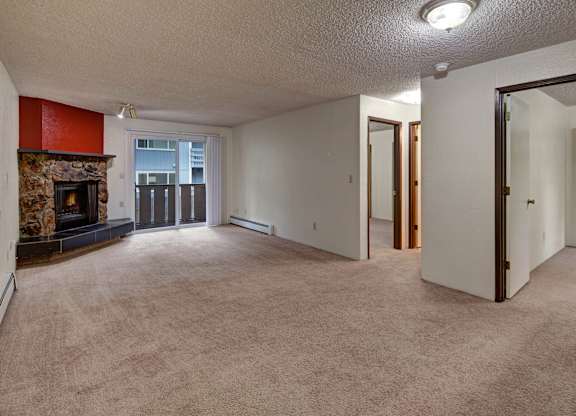 A spacious and inviting living room here at The Greenbriar featuring soft wall-to-wall carpeting, neutral walls, and a cozy stone fireplace with a bold accent surround. The room includes sliding glass doors leading to a private balcony, multiple doorways for easy flow to bedrooms and closets, and overhead lighting that creates a warm, comfortable atmosphere.