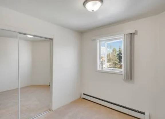 A bedroom here at La Maisonnette with beige carpet and white walls. A window with vertical blinds brings in natural light, and mirrored closet doors provide storage and reflect the space.