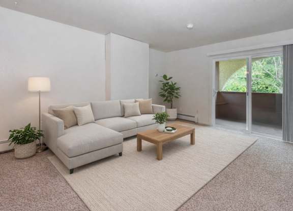 A living room here at Ladera Villa, with beige carpet and white walls. A light-colored sectional sofa sits on an area rug with a wooden coffee table, and a sliding glass door opens to a private balcony with greenery outside.