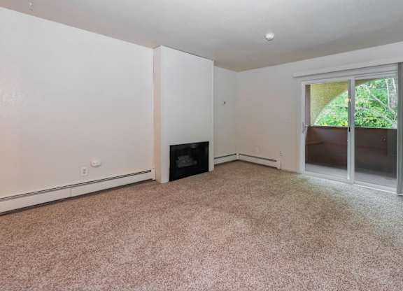 An open-concept and empty living room here at Ladera Villa, with beige carpet and neutral walls. A fireplace is set into the wall, and a sliding glass door opens to a private balcony with greenery outside, creating a welcoming atmosphere.