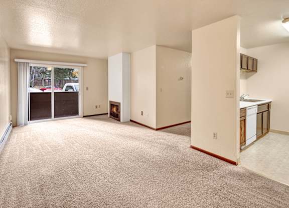 A spacious, empty living room here at Ladera Villa, with beige carpet and neutral walls. A fireplace adds warmth, and a sliding glass door opens to a private balcony, with the kitchen visible on the right-hand side.