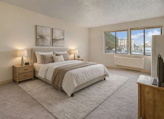 A bright, cozy bedroom here at Mendenhall-Tower with soft neutral carpet, a comfortable bed dressed in white linens, and warm bedside lighting. A large window brings in natural light and scenic city views, creating an airy and relaxing atmosphere.
