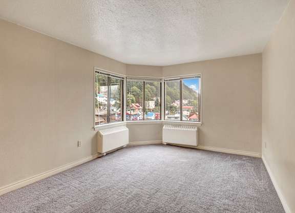 A bright, spacious corner room here at Mendenhall-Tower with soft gray carpeting, neutral walls, and large wraparound windows showcasing scenic hillside views and abundant natural light.