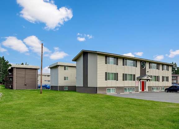 A three-story Montclair apartment building with a neutral exterior, a bright red entry door, and open green lawn space under a sunny blue sky.