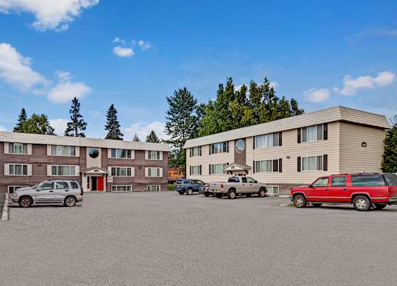 A wide exterior view of Montclair showing multiple three-story apartment buildings with neutral siding, red entry doors, and an open parking lot surrounded by mature trees.