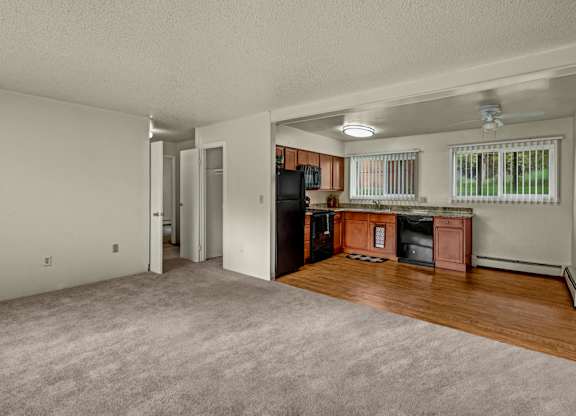 A spacious open-concept living area here at Montclair with plush beige carpet, an adjoining kitchen with warm wood cabinets and black appliances, and large windows with vertical blinds for natural light.